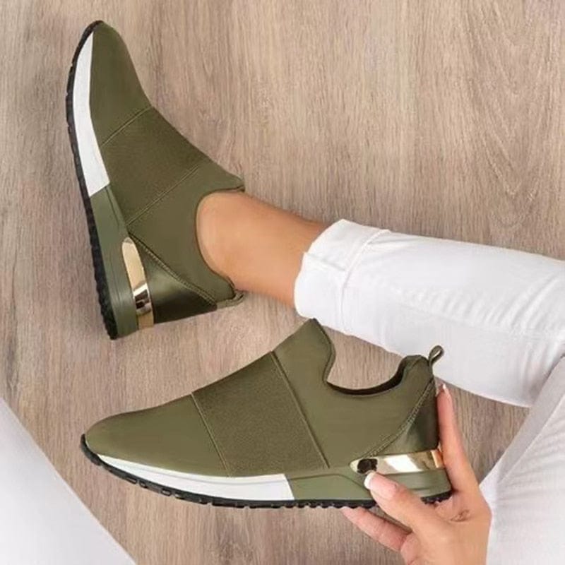 Olijfgroene slip-on sneakers met witte zool en gouden details, gedragen door persoon in witte broek op houten vloer. Comfortabele damesmode schoenen.